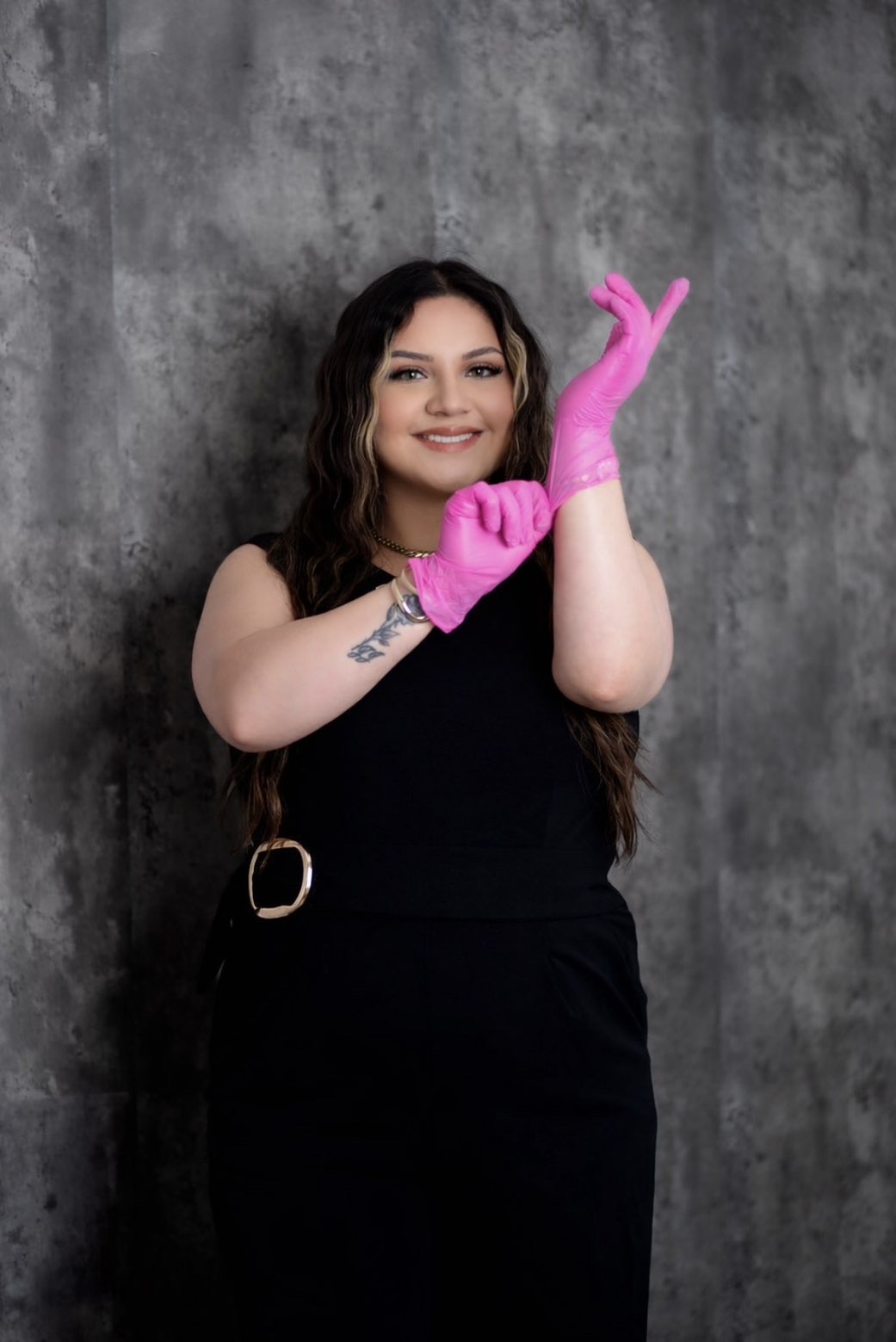 Moni smiling in a black jumpsuit, pulling on pink gloves in front of a grey backdrop.