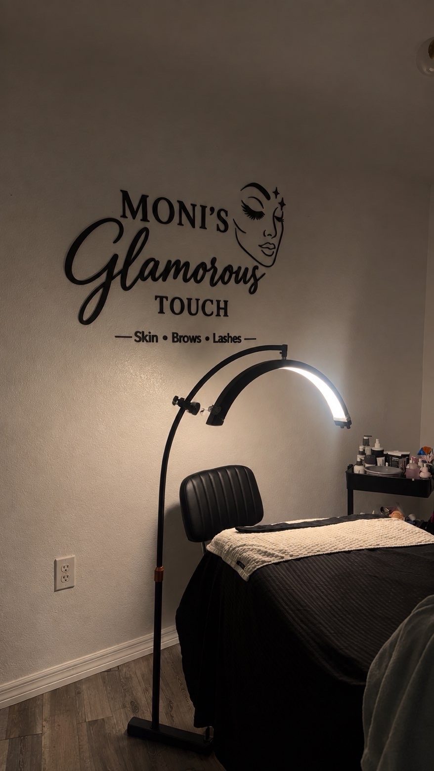 Inside the studio: the black Moni's Glamorous Touch sign, reading Skin, Brows, Lashes, on the wall above a treatment bed, with an arched lash lamp and a cart of skincare products.